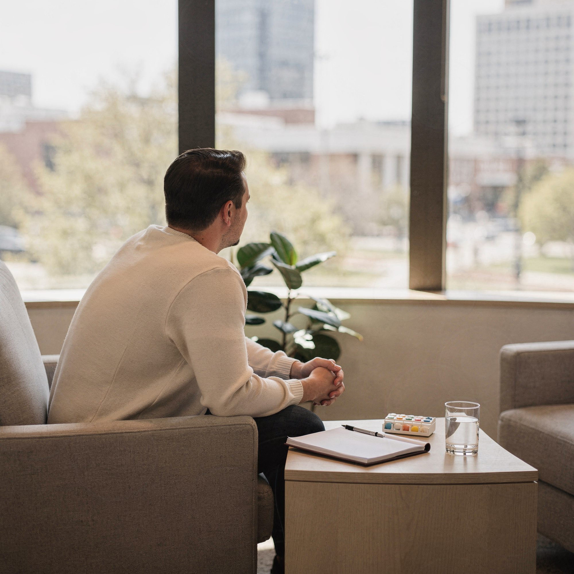 Man seated in a modern clinical waiting area with city skyline view, medication organizer and notepad on table before him